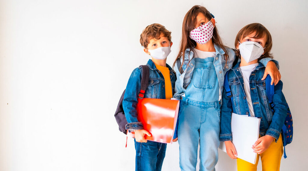 Niños con mascarillas sosteniendo sus pertenencias escolares y de pie contra una pared