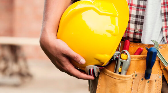 Primer plano de casco de constructor sostenido por albañil. También se observa un bolsillo con herramientas.