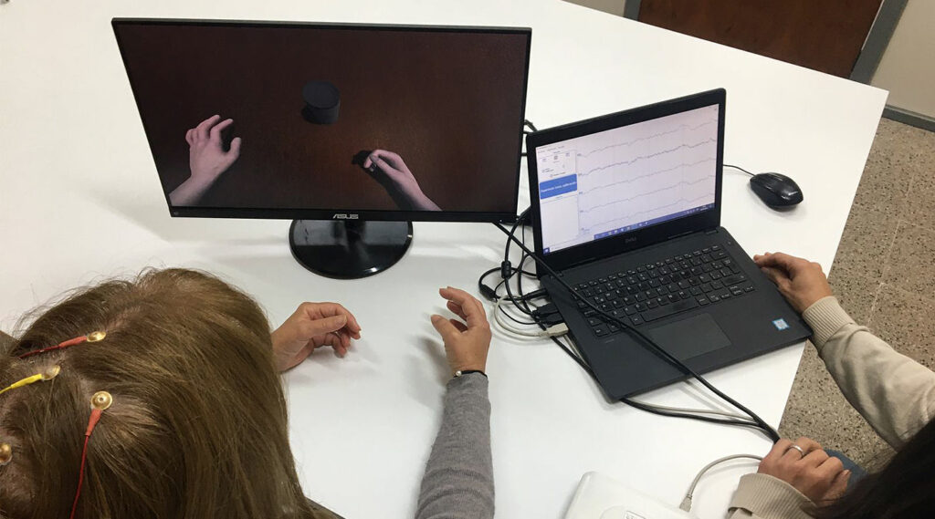 Dos mujeres y dos computadoras; se muestra en una pantalla la actividad eléctrica que miden electrodos en la cabeza de una de las mujeres, y en la otra computadora se observa en primera persona, brazos y manos en un espacio tridimensional con objetos.