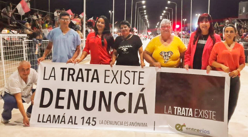 Referentes del Consejo Provincial de Prevención, Protección y Asistencia a las Víctimas y Testigos de la Trata y el Tráfico de Personas del Ministerio de Gobierno y Justicia posando junto a una bandera contra la trata de personas en un carnaval provincial.