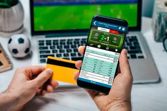 Notebook con un partido de fútbol de fondo y primer plano de mano sosteniendo smartphone con un sitio de apuestas on line.