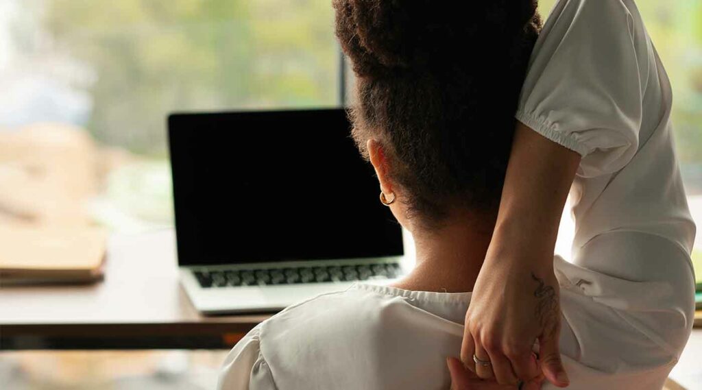 Mujer sentada de espaldas frente a su notebook tomándose el cuello, dolorida.