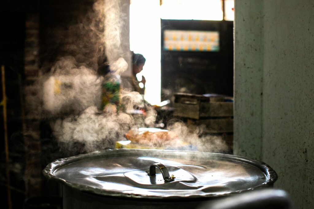 En una casa humilde se ve en primer plano una olla de aluminio con la tapa abollada humeando, y de fondo una mujer barriendo